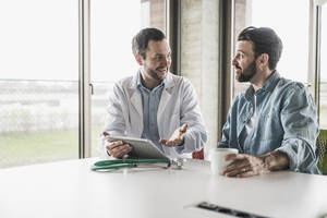 Lächelnder Arzt, der einen Tablet-PC neben einem am Schreibtisch sitzenden Patienten in einer Klinik hält - UUF28608