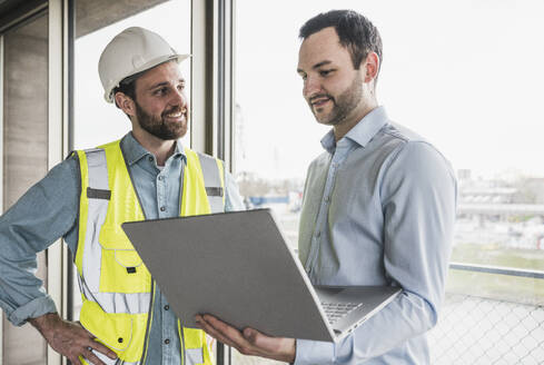 Lächelnder Bauunternehmer mit Blick auf einen Architekten mit Laptop - UUF28617