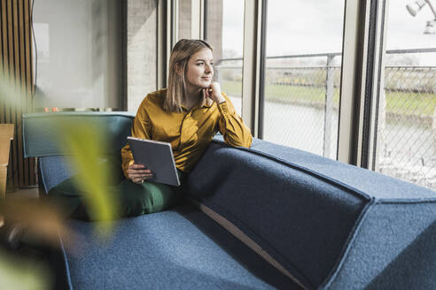Nachdenkliche Geschäftsfrau, die einen Tablet-PC hält und sich auf eine Couch im Büro lehnt - UUF28636