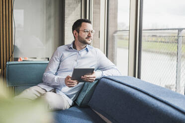 Geschäftsmann mit Brille lehnt auf der Couch und hält einen Tablet-PC im Büro - UUF28638