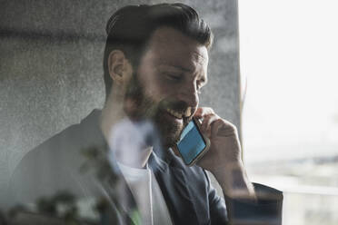 Lächelnder Geschäftsmann, der mit seinem Smartphone im Büro spricht, gesehen durch Glas - UUF28641