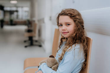 Porträt eines kleinen Mädchens mit einem Teddybär, das im Krankenhausflur sitzt und wartet. - HPIF09917