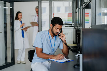 Junger Krankenpfleger, der am Empfang sitzt und telefoniert, im Hintergrund diskutieren seine Kollegen. - HPIF09943
