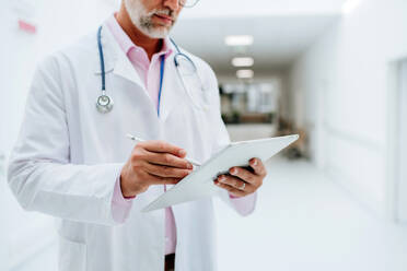 Mittelteil eines reifen Arztes mit digitalem Tablet in einem Krankenhausflur. - HPIF09956