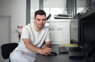 Junger Mann mit Down-Syndrom arbeitet in einem Krankenhausbüro und schreibt etwas am Computer. Konzept der Integration von Menschen mit Behinderungen in die Gesellschaft. - HPIF10149