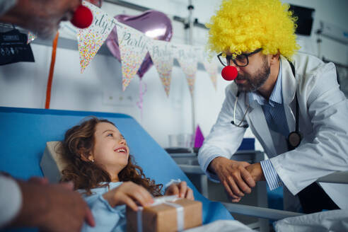 Glücklicher Arzt mit roten Clownsnasen feiert Geburtstag mit kleinem Mädchen in einem Krankenhauszimmer. - HPIF10240