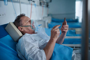 Senior man with oxygen mask lying in the hospital bed and scrolling smartphone. - HPIF10247