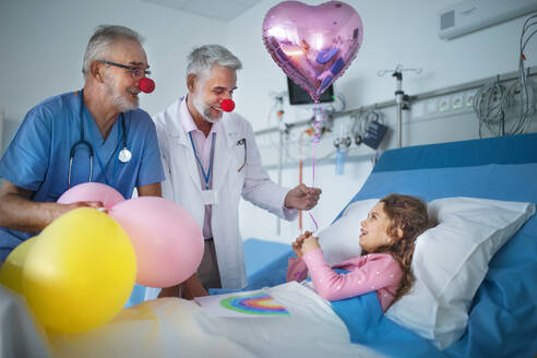 Glückliche Ärzte mit roten Clownsnasen feiern Geburtstag mit einem kleinen Mädchen in einem Krankenhauszimmer. - HPIF10284