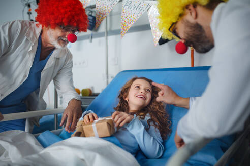 Glückliche Ärzte mit roten Clownsnasen feiern Geburtstag mit einem kleinen Mädchen in einem Krankenhauszimmer. - HPIF10303