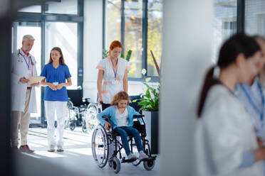 Junge Krankenschwester schiebt kleines Mädchen im Rollstuhl auf einem Krankenhausflur. - HPIF10321