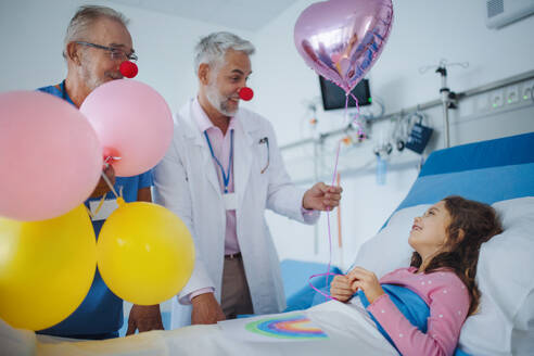 Glückliche Ärzte mit roten Clownsnasen feiern Geburtstag mit einem kleinen Mädchen in einem Krankenhauszimmer. - HPIF10354