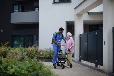 Pflegekraft geht mit einer älteren Kundin vor einem Pflegeheim spazieren. - HPIF10416