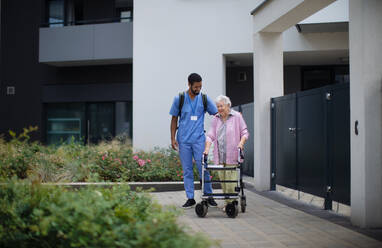 Pflegekraft geht mit einer älteren Kundin vor einem Pflegeheim spazieren. - HPIF10417