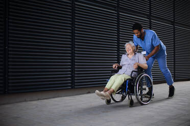 Ein pflegender Mann schiebt eine aufgeregte ältere Frau im Rollstuhl auf einer Straße und genießt die gemeinsame Zeit. - HPIF10418