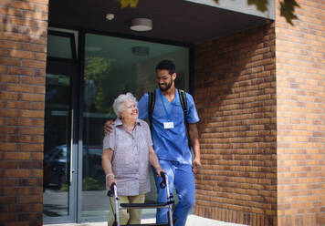 Pflegekraft geht mit einer älteren Kundin vor einem Pflegeheim spazieren. - HPIF10425