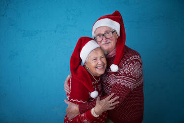 Porträt eines glücklichen älteren Paares in Weihnachtspullovern, das sich vor einem blauen Hintergrund umarmt. - HPIF10516