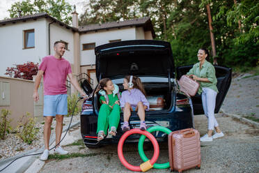 Ein glücklicher Vater hält ein Stromkabel in der Hand und lädt sein Elektroauto auf, der Rest der Familie packt die Koffer in den Kofferraum und bereitet sich auf den Urlaub vor. - HPIF10717