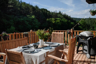 Ein Esstisch mit Holzstühlen für das Abendessen auf der Terrasse mit Grill im Sommer, Gartenparty. Konzept. - HPIF10733