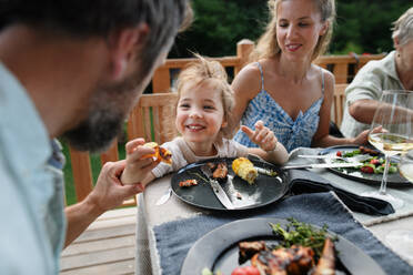 A family eating at barbecue party dinner on patio, little girl with parents sitting at table and enjoying food. - HPIF10745