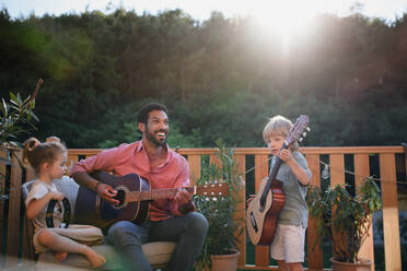 A young African american man enjoys playing the guitar with little children accompaining him with instruments on patio in garden in summer. - HPIF10759