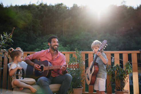 Ein junger afroamerikanischer Mann genießt das Gitarrenspiel mit kleinen Kindern, die ihn im Sommer auf einer Terrasse im Garten mit Instrumenten begleiten. - HPIF10759