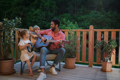 Ein junger afroamerikanischer Mann genießt das Gitarrenspiel mit kleinen Kindern, die ihn im Sommer auf einer Terrasse im Garten mit Instrumenten begleiten. - HPIF10760