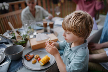 Ein kleiner Junge isst gebratenen Mais und Gemüse bei einem Familiengrillfest im Garten. - HPIF10775