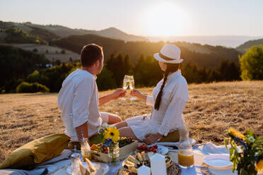 Ein attraktives, verliebtes Paar genießt ein Picknick und trinkt Wein auf einem Hügel bei Sonnenuntergang. - HPIF10860