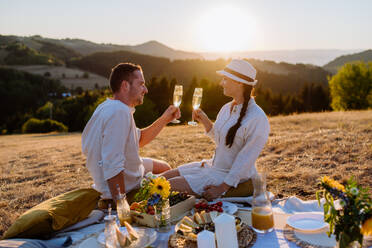 Ein attraktives, verliebtes Paar genießt ein Picknick und trinkt Wein auf einem Hügel bei Sonnenuntergang. - HPIF10861