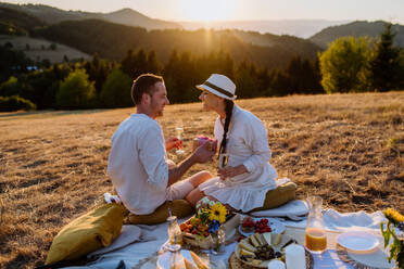 Ein attraktives, verliebtes Paar genießt ein Picknick und trinkt Wein auf einem Hügel bei Sonnenuntergang. - HPIF10862