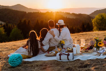 Familie mit Kindern beim Picknick in der Natur mit Blick auf den Sonnenuntergang, Rückansicht. - HPIF10868