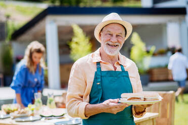 Ein glücklicher älterer Mann serviert Burger auf einem Mehrgenerationen-Gartenfest im Sommer. - HPIF10896