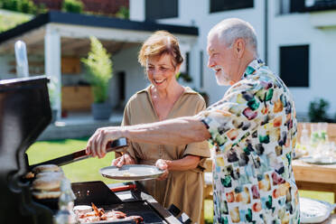 Ein älterer Mann grillt draußen im Garten und gibt seiner Frau einen frischen Hamburger. - HPIF10910