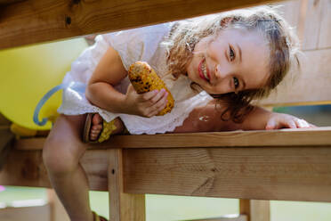 Ein süßes kleines Mädchen isst gegrillten Mais und schaut unter dem Tisch in die Kamera während einer Gartenparty. - HPIF10917