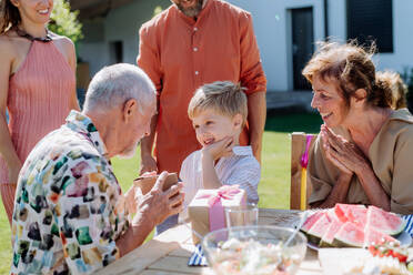 A happy little boy giving birthday present to his senior grandfather at generation family birthday party in summer garden - HPIF10923