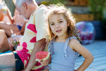 Cute little girl in a swimsuit eating watermelon corn and sitting next to pool with family at garden party. - HPIF10933