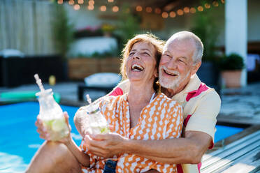 A happy senior couple enjoying drinks when relaxing and sitting by swimming pool in summer. - HPIF10947