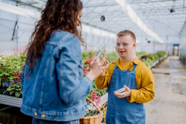 Ein glücklicher junger Angestellter mit Down-Syndrom arbeitet in einem Gartencenter und nimmt die Bezahlung von einem Kunden entgegen. - HPIF11015
