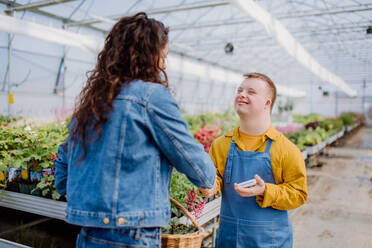 Ein glücklicher junger Angestellter mit Down-Syndrom arbeitet in einem Gartencenter und nimmt die Bezahlung von einem Kunden entgegen. - HPIF11016