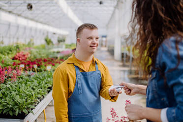 Ein glücklicher junger Angestellter mit Down-Syndrom arbeitet in einem Gartencenter und nimmt die Bezahlung von einem Kunden entgegen. - HPIF11017