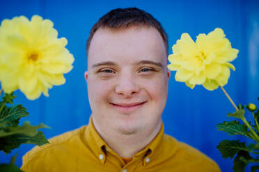 Ein glücklicher junger Mann mit Down-Syndrom schaut in die Kamera und hält Topfblumen vor blauem Hintergrund. - HPIF11050