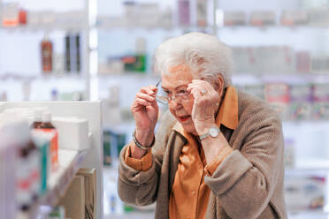 Senior woman reading text on the medication box, standing in pharmacy store. - HPIF11077