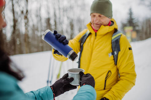 Ein älteres Ehepaar ruht sich mitten im verschneiten Wald aus und trinkt heißen Tee aus einer Thermoskanne. - HPIF11209