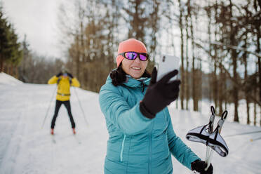 Senior woman taking selfie with her husband in background during cross country skiing in snowy nature. - HPIF11313