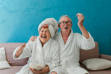Ein glückliches älteres Ehepaar sitzt im Bademantel auf einem Sofa und sieht mit Popcorn fern. - HPIF11346