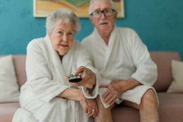 Ein glückliches älteres Paar sitzt im Bademantel auf einem Sofa und sieht fern. - HPIF11420