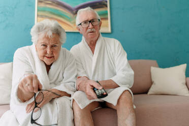 Happy senior couple sitting at a sofa in bathrobes and watching TV with popcorn. - HPIF11422