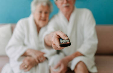 Ein glückliches älteres Paar sitzt im Bademantel auf einem Sofa und sieht fern. - HPIF11423