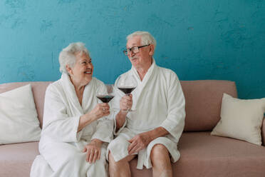 Happy senior couple sitting together in the bathrobe on sofa with glass of wine, having nice time at home. - HPIF11424