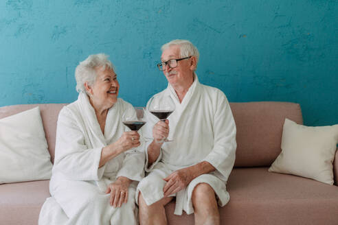 Glückliches älteres Paar, das zusammen im Bademantel auf dem Sofa mit einem Glas Wein sitzt und eine schöne Zeit zu Hause hat. - HPIF11424